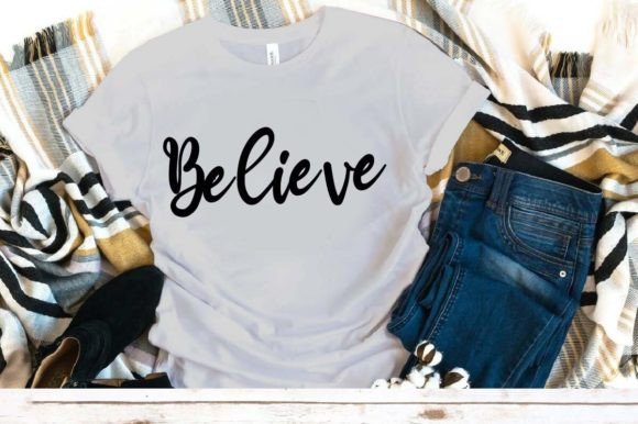 White t-shirt with 'Believe' in black script, laid on a plaid blanket with jeans and black shoes.