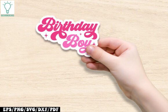 A hand holding a pink and white sticker with the phrase 'Birthday Boy' in a groovy font on a wooden surface.