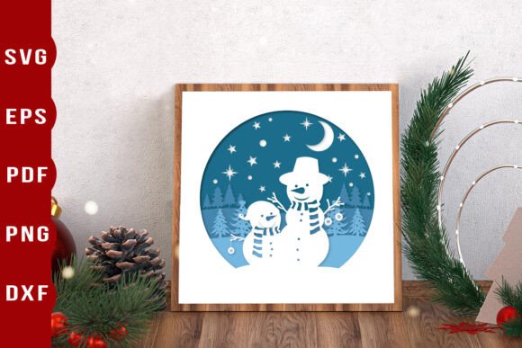 Framed paper cut design of two snowmen with scarves under a starry sky and crescent moon.