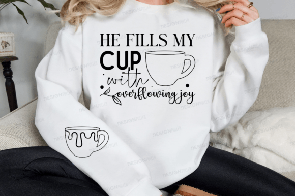 Person seated in a white sweatshirt featuring 'He Fills My Cup with Overflowing Joy' and mug illustrations.