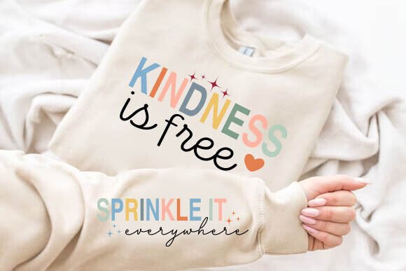 Beige sweatshirt with 'Kindness is free' in colorful letters and a heart, sleeve says 'Sprinkle it everywhere'.