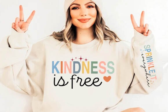 Woman wearing beige sweatshirt with 'Kindness is free' design, sleeve reads 'Sprinkle it everywhere' vertically.